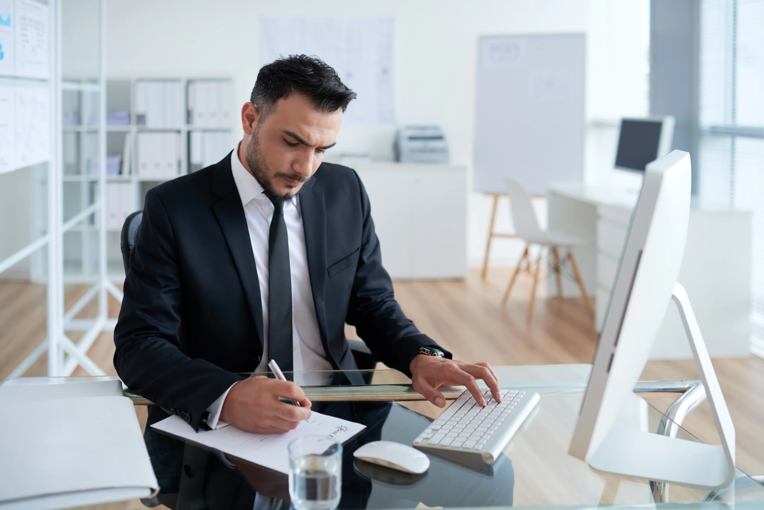 caucasian-man-business-suit-sitting-office-working-computer-writing-paper